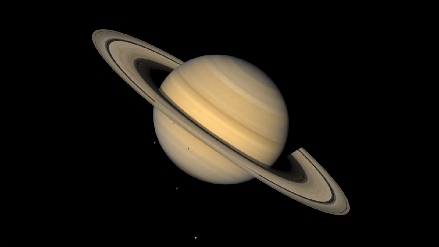 This image shows Saturn, ringed and in tones of tan, against an inky background. Three tiny light spots in the background and one dot against the lower half of the planet are four of Saturn's moons.