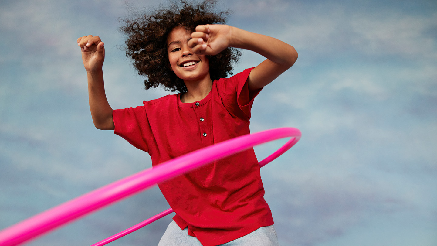 A smiling child swings a pink hula hoop around their waist.