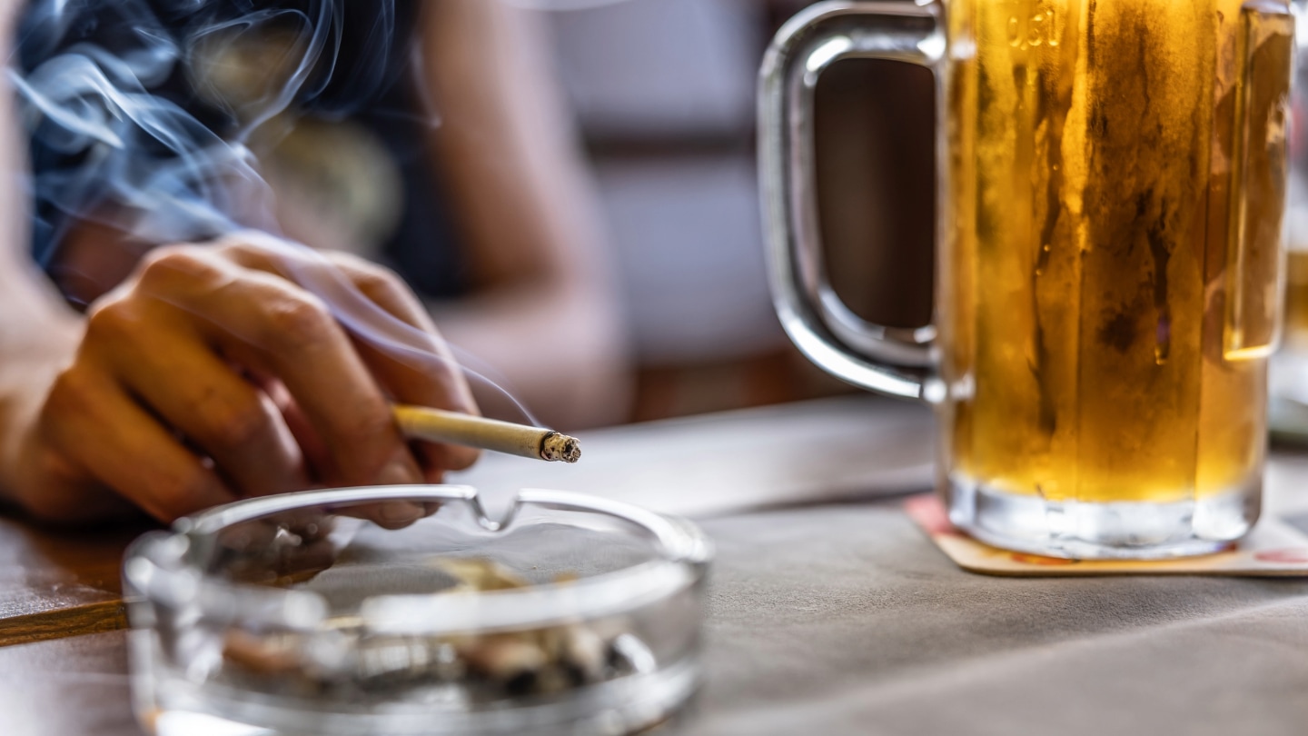 A person's hand holding a lit cigarette near an ashtray and a mug of peer.