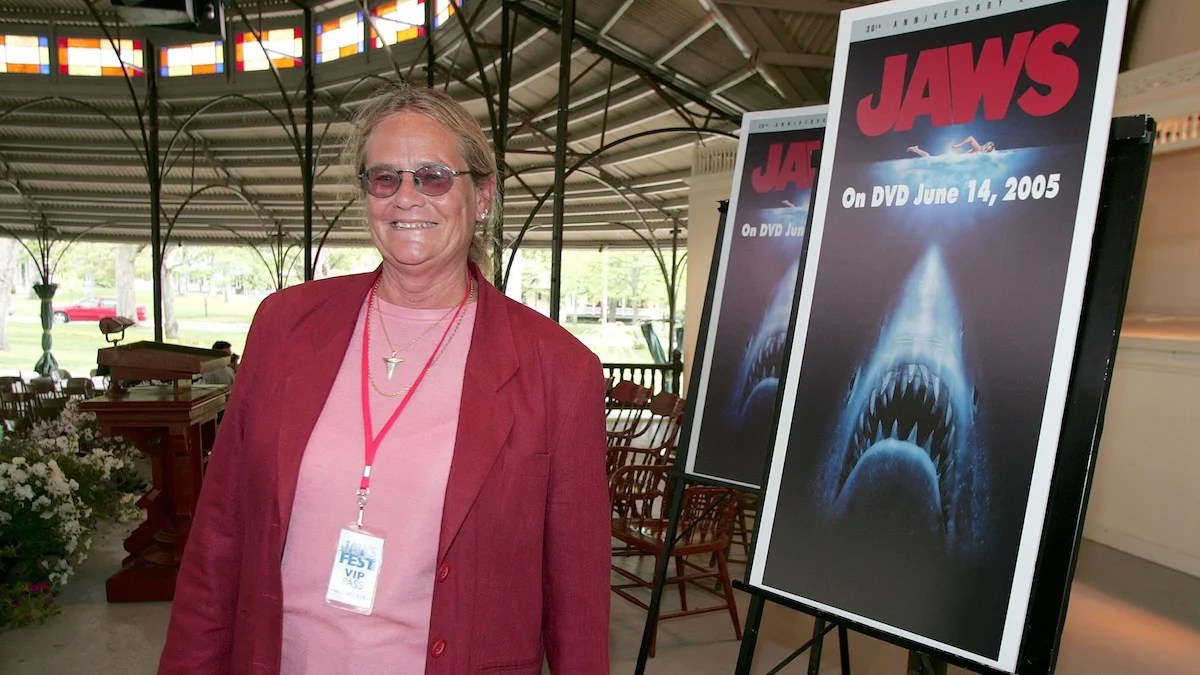 An older woman stands in a covered area next to a poster for Jaws on DVD. She has light-toned skin and blond hair.