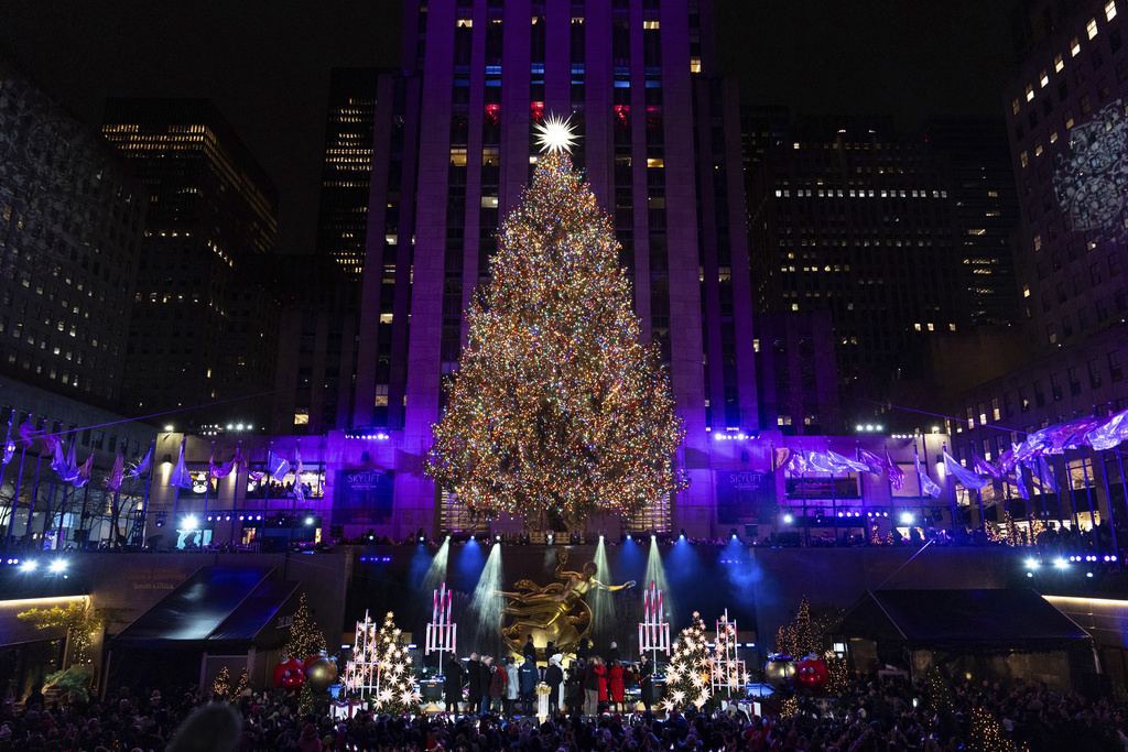 A large evergreen lit with colorful holiday lights and a star amidst a city landscape.