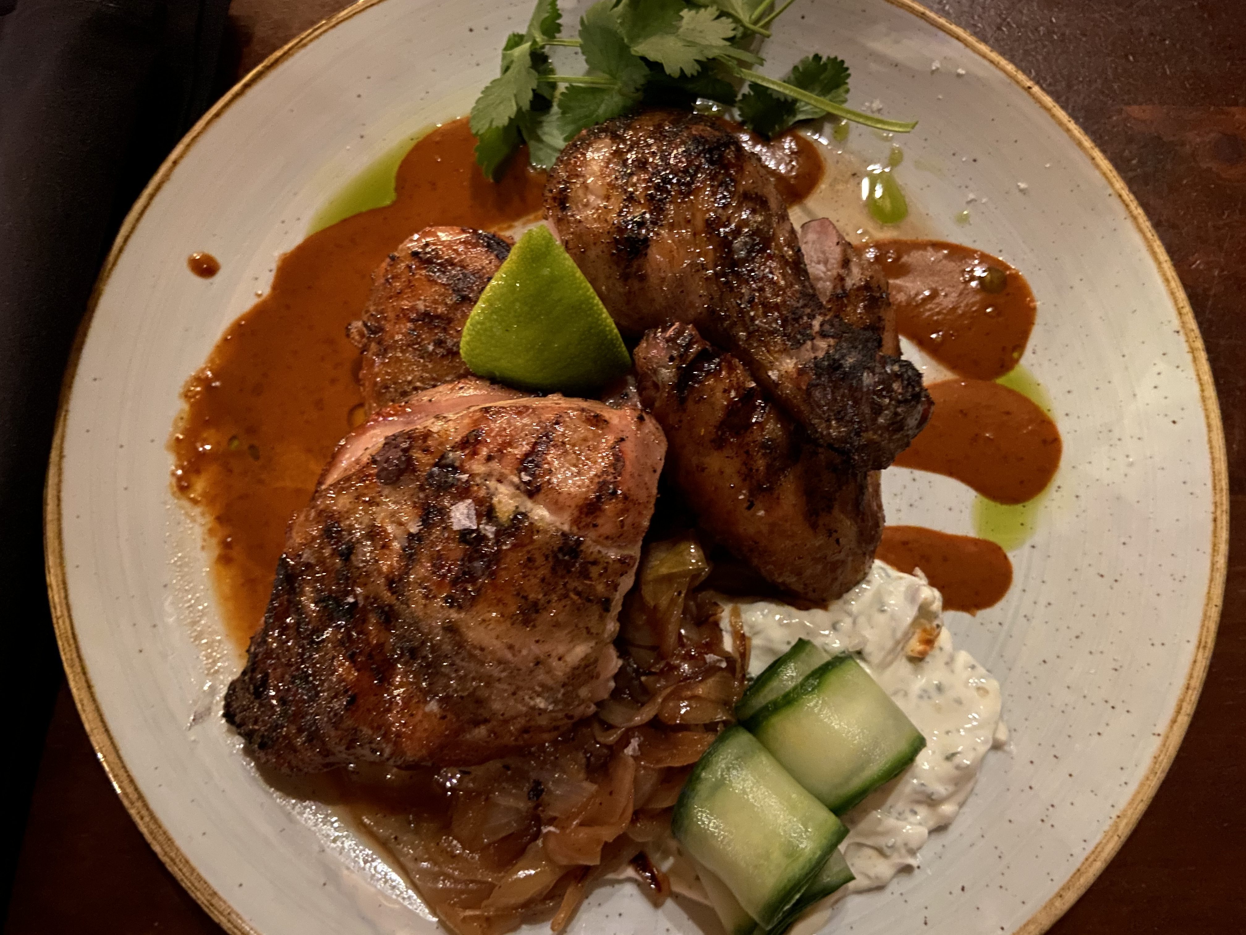 an overhead photo of a crispy grilled half chicken with a brick-red sauce, and some rolled ccumber slices and labneh