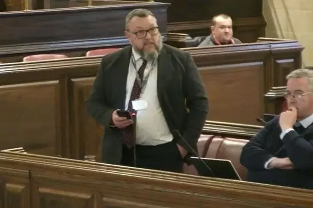 Man with beard and glasses in suit stood up at council chamber benches giving a speech