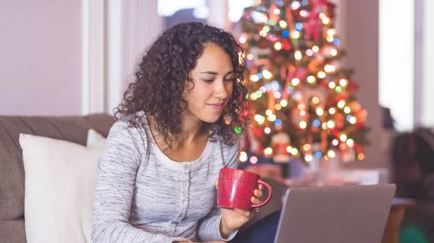 A young woman cozies up on the couch in front of her computer and holds a credit card and cup of tea in the other.