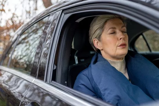 A medium shot of a woman who has recently recovered from cancer. She takes a moment of reflection and anxiety before leaving the car for her follow-up appointment. Dressed warmly in a navy blue coat, she is mentally preparing herself for the check-up. The image captures the emotional weight of her journey and the uncertainty that accompanies post-recovery medical visits.