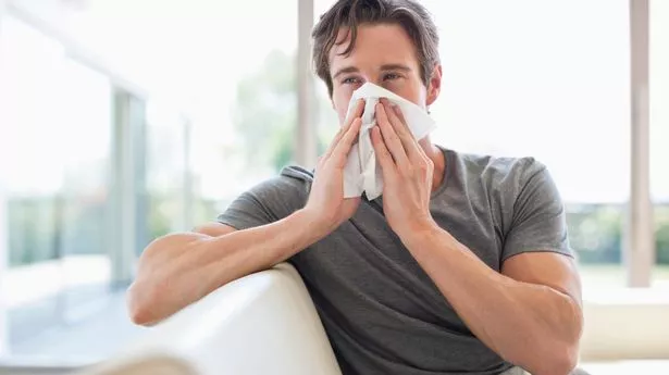 Man in pjs sits on sofa sneezing into tissue