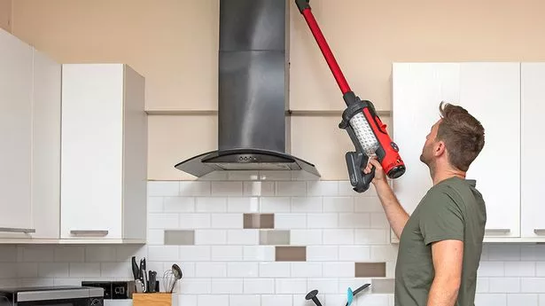 Man in green t-shirt and blue denim jeans uses red and black hoover to clean top of oven in white and brown kitchen