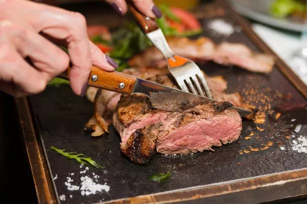 Woman hand holding knife and fork cutting grilled beef steak on stoned plate. Selective focus