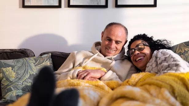 A shot of a mature heterosexual couple laying on the sofa together with a blanket over them. They are staying warm during the cost of living crisis.