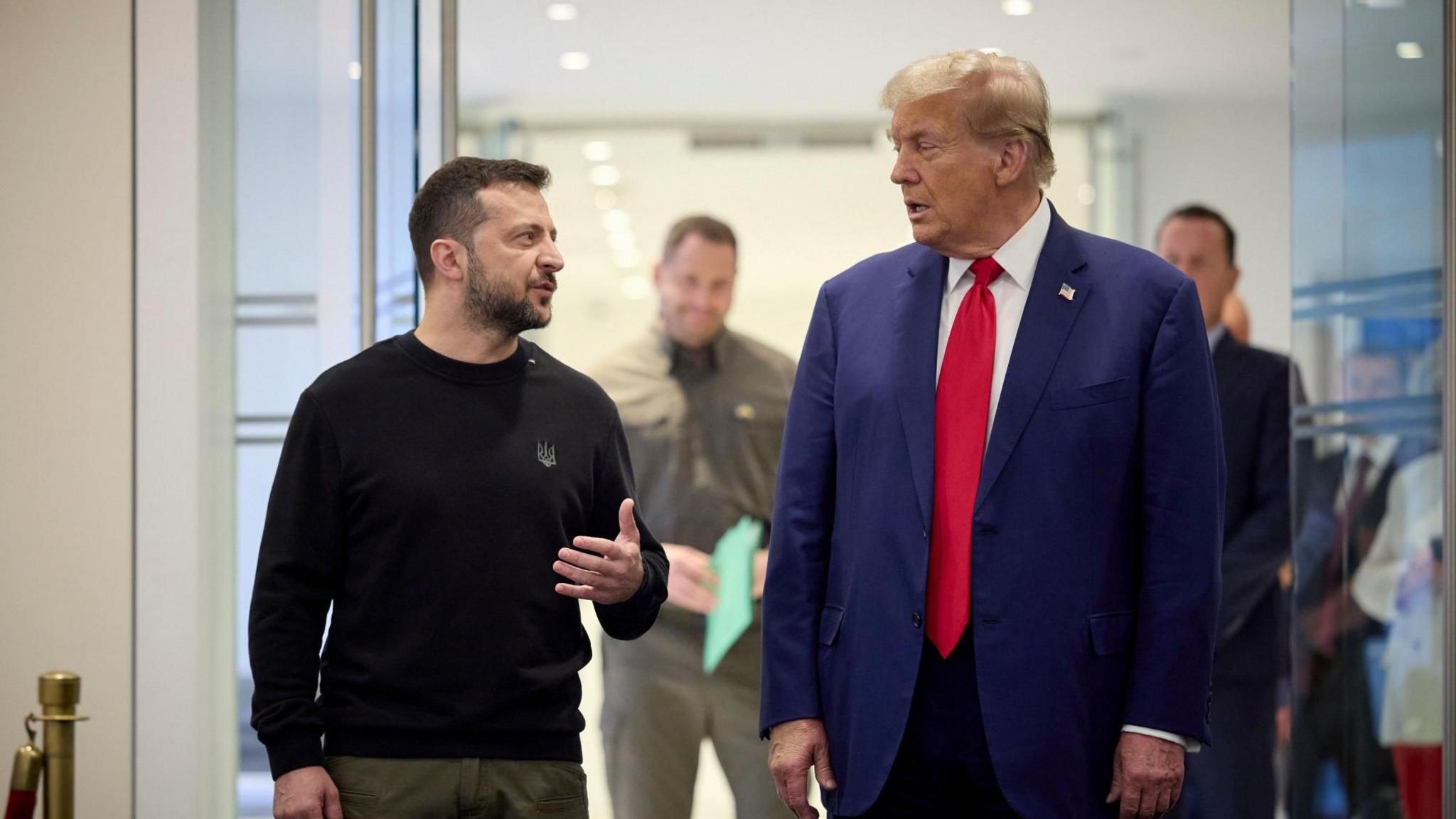 Ukrainian President Volodymyr Zelensky, dressed in a black sweater with a Ukrainian coat of arms emblem, engages in conversation with President-elect Donald Trump, wearing a blue suit, white shirt, and bright red tie.