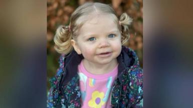 A young girl with blue eyes and blond hair tied up in a pig tail. She is wearing a colourful jacket and a pick jumper with flowers on