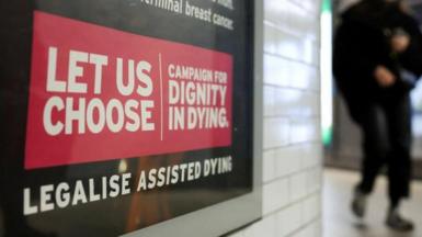 A Tube passenger walks past an assisted-dying rights billboard ahead of the parliamentary debate on Friday, in London, Britain November 26, 2024. The sign says 'Let us choose - campaign for dignity in dying' in white letter against a red background