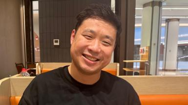 Sun Zhan, 25, smiles and tilts his head to the side as he poses for a picture inside what looks like a restaurant. 