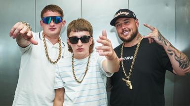 Sam Robinson (Clive), Gareth Kelly and Kane Welsh from Bad Boy Chiller Crew pose, with their fingers doing salute signs. The trio are wearing gold chains around their necks, with Clive and Kane also wearing sunglasses and pale tshirts. Gareth is wearing a black tshirt and cap.