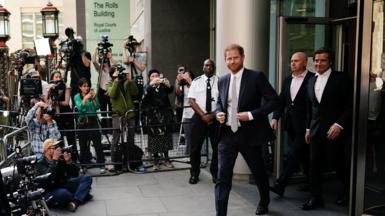 Prince Harry leaves the court during his hacking case against the Daily Mirror. He wears a dark coloured suit, white shirt and tie. His barrister David Sherborne, also dressed in a dark suit is on his left.  A crowd of photographers are behind a metal barrier