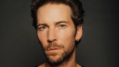 An artistically lit headshot of a man in front of a dark background. He's got a neutral expression, a silver hoop earring in his left ear and his angular chin is framed by a short beard.