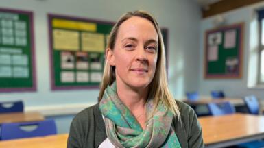 A head and shoulders photo of a woman with straight blonde hair looking at the camera. She is wearing a green paisley scarf, olive cardigan and white blouse. She is sitting in a school classroom with wooden tables and blue chairs behind her, as well as display boards against the back wall.