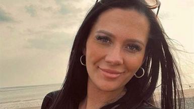 Kiena Dawes, with long dark hair, long eyelashes and silver hoop earrings, smiles at the camera in front of a beach