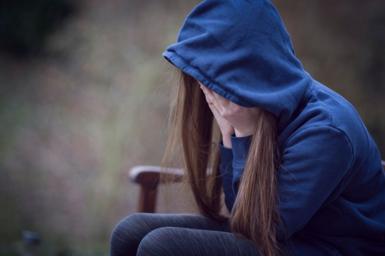 A young girl with her head in her hands, wearing a blue hoodie that obscures her identity