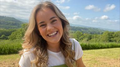 Heledd Roberts smiling as she looks at the camera. Behind her, you see rolling hills and a blue sky in what is a picturesque location. There are tree and green areas scattered around the background suggesting Heledd is in a rural location.