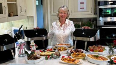 Beverley Jarvis, wearing a white lace shirt, is standing behind her kitchen island, with her hands placed on two large air fryers, with another air fryer at the sides and plates and dishes of Christmas food and small Christmas decorations in front of her. Behind her are cream-coloured traditional kitchen wall cabinets and an oven.