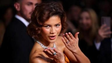 Actress Zendaya smiles and blows a kiss to someone off camera as she walks the red carpet at the Golden Globes. On the fourth finger of her left hand is a large diamond ring, not normally seen there, suggesting she has got engaged. She wears a burnt orange dress with shoulders exposed, and a large diamond necklace.