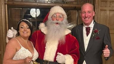 Lucy and Lee Holliday pose and smile in their wedding outfits either side of Father Christmas who arrived for their wedding reception