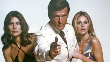 Sir Roger Moore, with actresses Maud Adams and Britt Ekland, on the set of the James Bond film, The Man With the Golden Gun. Sir Roger is wearing a white jacket and black tie and is pointing a gun down the lens of the camera. Maud and Britt are dressed in swimwear and are staring directly at the camera.