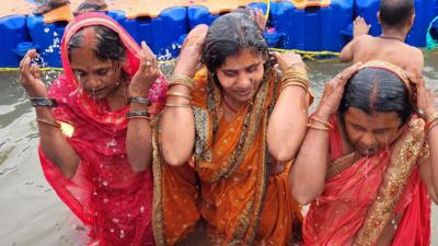 Three women wearing sarees and gold bangles taking a dip in the sacred river during the Mahakumbh Mela in India's Prayagraj city