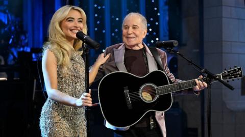 Sabrina Carpenter, with blonde curled hair, smiles holding a mic and Paul Simon's shoulder as he looks towards her wearing a velvet blazer and holding a black acoustic guitar