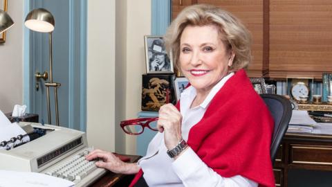 Barbara Taylor Bradford sitting at a typewriter in an office and smiling at the camera