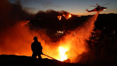 A firefighter uses a hose while a helicopter dumps water on a blazing fire in Los Angeles