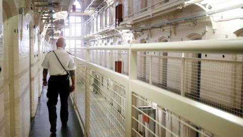 Inside Barlinnie, a central area with cells on either side of a hallway.  A guard is walking away from the camera down the hall. 