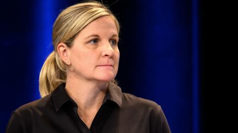 Kirsty Coventry, with her blonde hair tidied behind her ears into a ponytail, wears a black blouse as she stands in front of a dark blue background