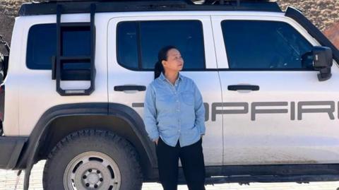 Su Min is standing in front of her SUV. She is wearing a blue shirt and black trousers and has her hands in the pockets