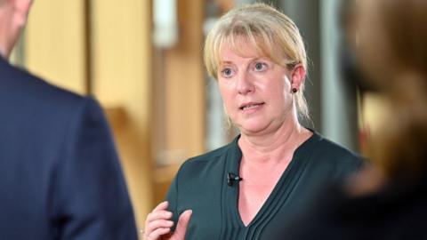 Shona Robison, with blonde hair and wearing a green dress, speaks to reporters in the Scottish Parliament 