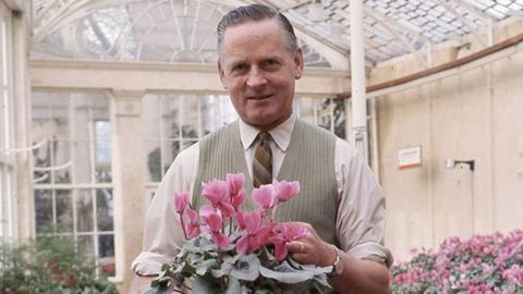 A man in a waitcoat and tie holding a plant with pink flowers