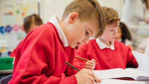 A primary school student writes in a book with a pencil while another student sits in the background.