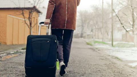 Man in sheepskin coat walks down a snowy path with houses and bare trees on either side. He is dragging a black suitcase.