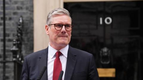 Sir Kier Starmer outside 10 Downing street. He's wearing a dark suit with a red tie and glasses. The no 10 door is gloss black with the number showing prominently.