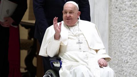 Pope Francis is waving to people as he is pushed in a wheelchair by someone assisting him.