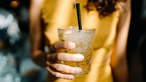 A close up photo shows a cocktail glass with golden coloured liquid inside, held by a hand outstretched. The drink has a straw and the owner of the drink has a yellow top, appears to be in a bar setting.