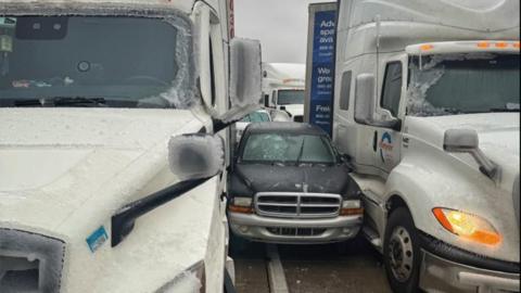 Black car stuck in between two larger trucks, all covered in snow and ice