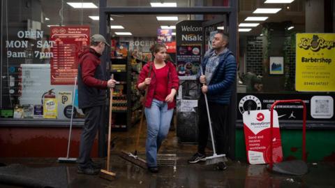 Staff with brooms clear water and mud from entrance to shop