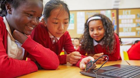 Three female choolchildren in red cardigans looking at a laptop. One of the children is holding a BBC micro:bit