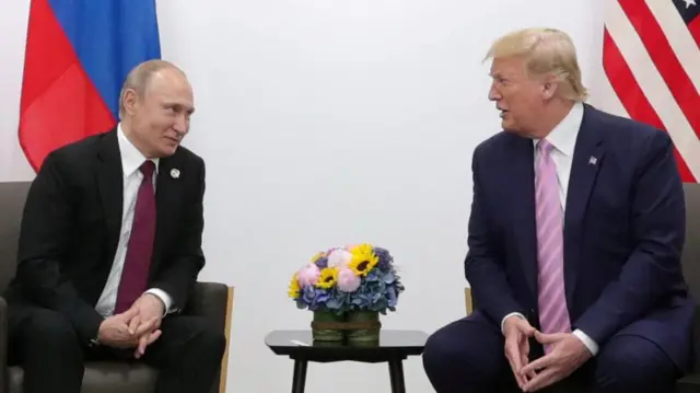 Valdimir Putin, the the left of the shot, looks across at Donald Trump. Both men are seated and there is a table with a small bunch of flowers on it in between them.