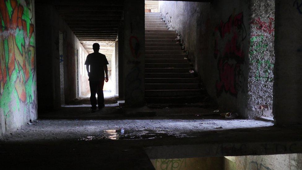 A lone figure walks down a dark hallway in the ruined building, with graffiti on the walls