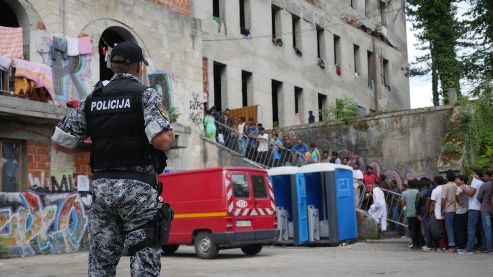 A police officer in a stab vest watches a long line of men on the stairway into the shell of a building