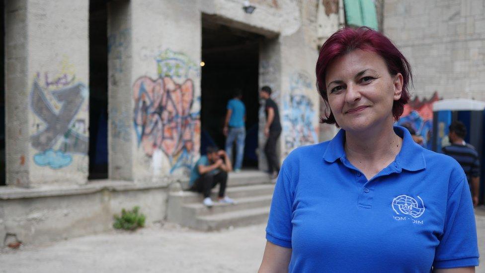 Amira, in a blue and white collared t-shirt, stands in front of graffiti-marked pillars between which men loiter