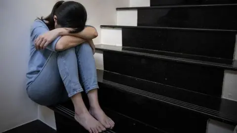 A barefoot woman wearing light blue jeans and a light blue top is sitting down on some stairs with her arms around her knees and her face in her lap. she is pale with dark brown hair in a ponytail